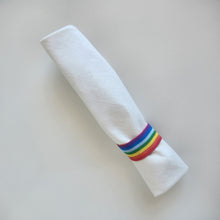 Load image into Gallery viewer, Rolled up cream white linen cutlery wrap secured with a rainbow stripe elastic on a light gray background