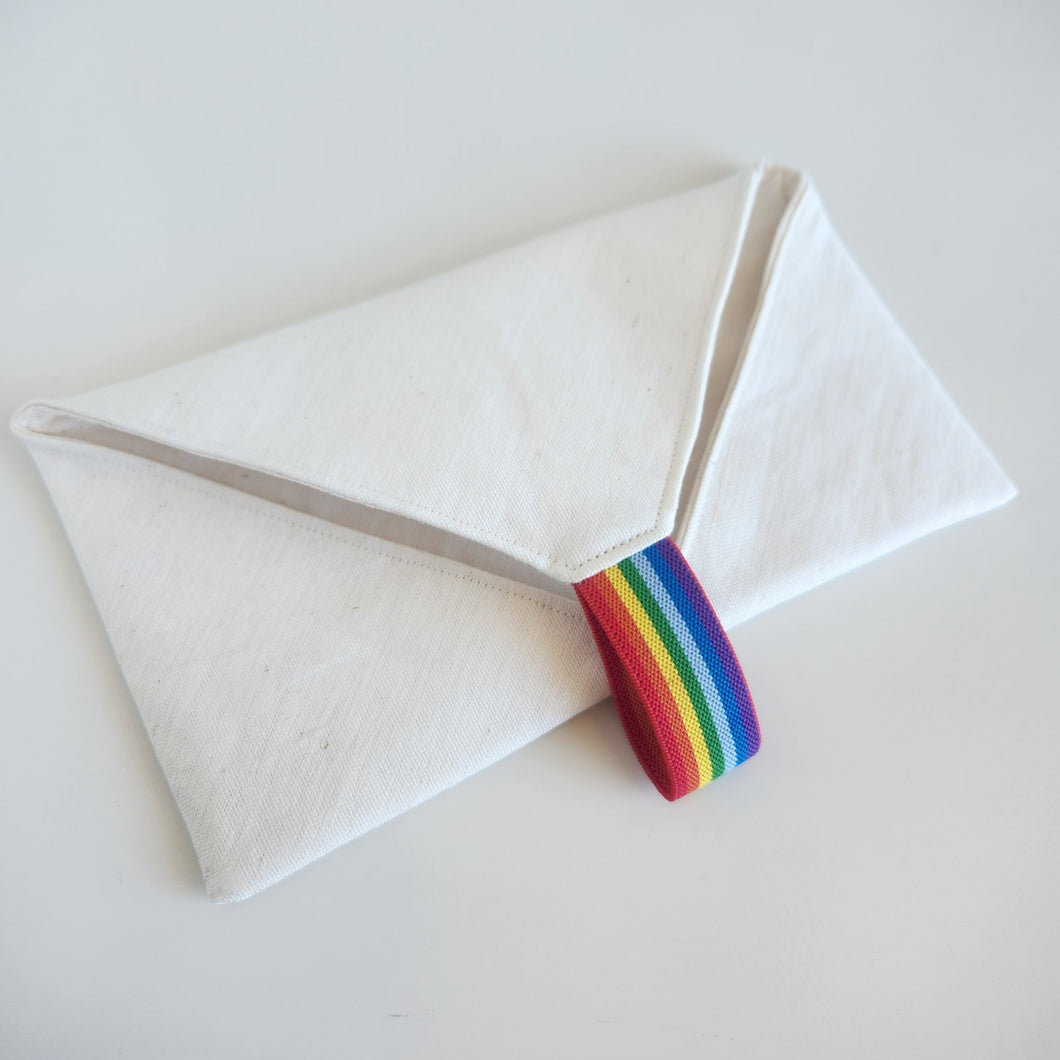 White linen cutlery wrap pouch with a rainbow striped elastic loop on a light gray background