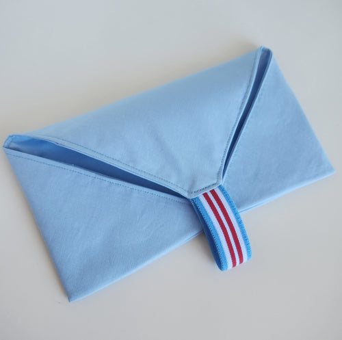 Folded light blue cotton fabric utensil cutlery wrap holder with a striped red, white, and blue elastic loop on a light gray background
